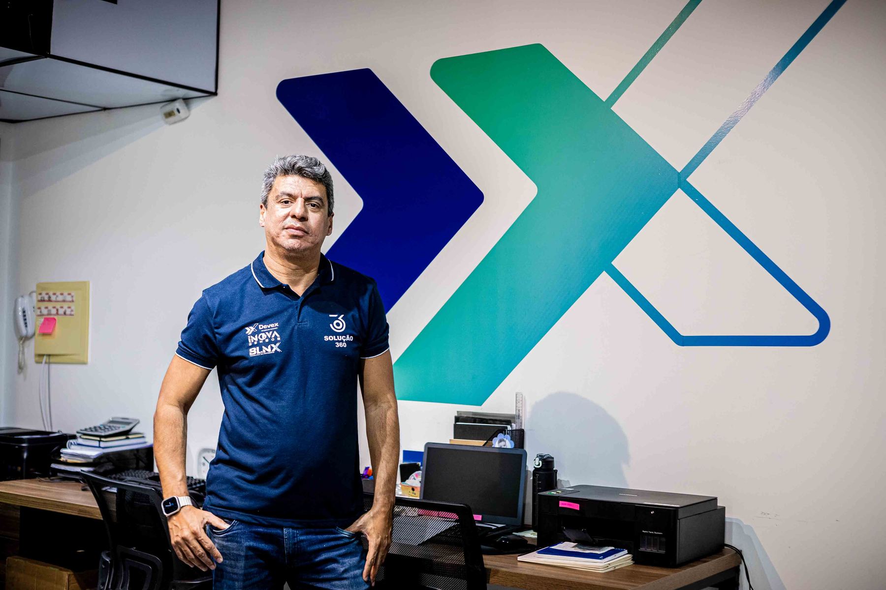 Líder da Devex de pé no escritório, diante da parede com o grande símbolo X da marca