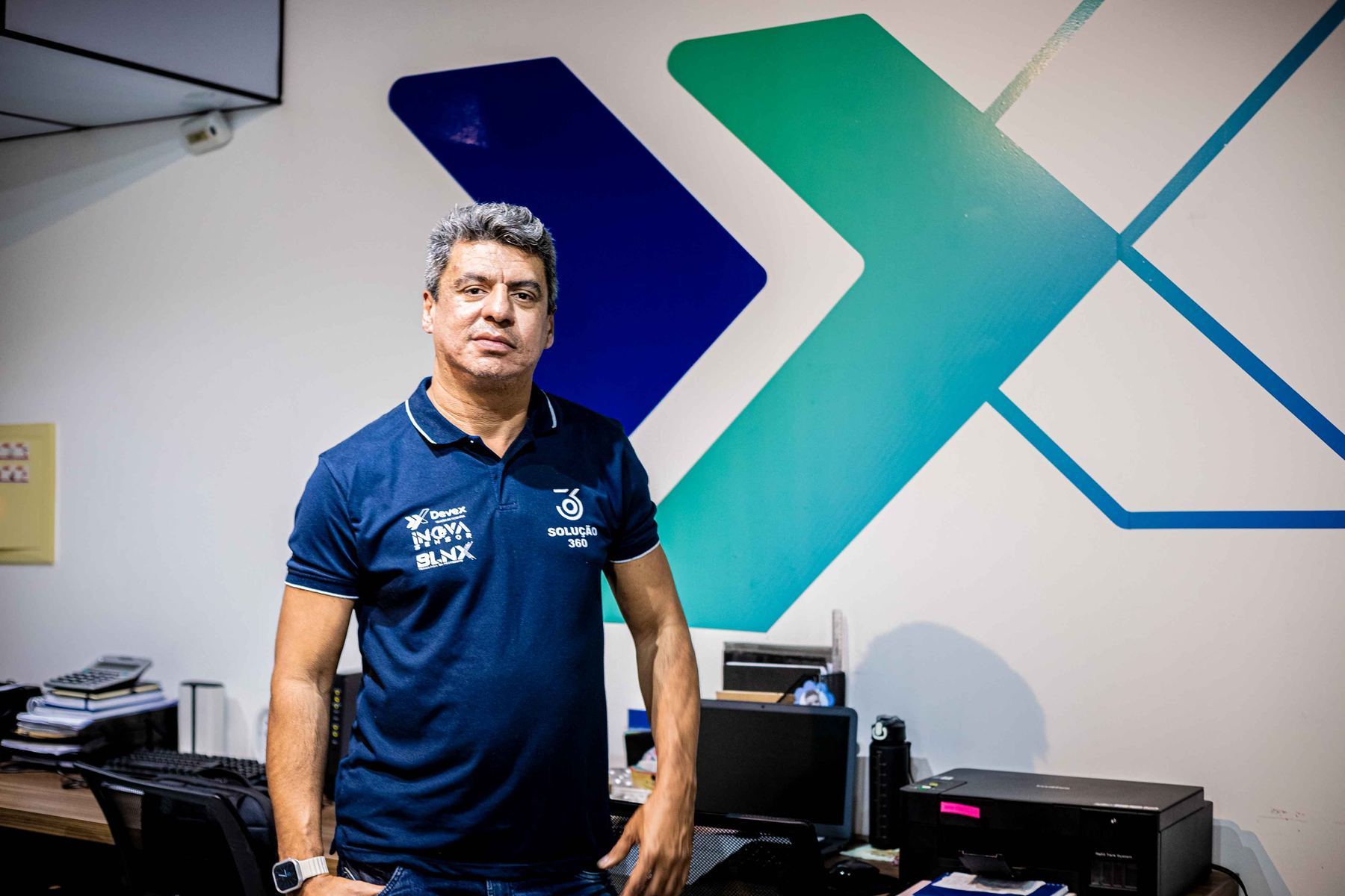Líder da Devex de camisa polo azul-marinho em frente à parede com o grande símbolo X da marca.