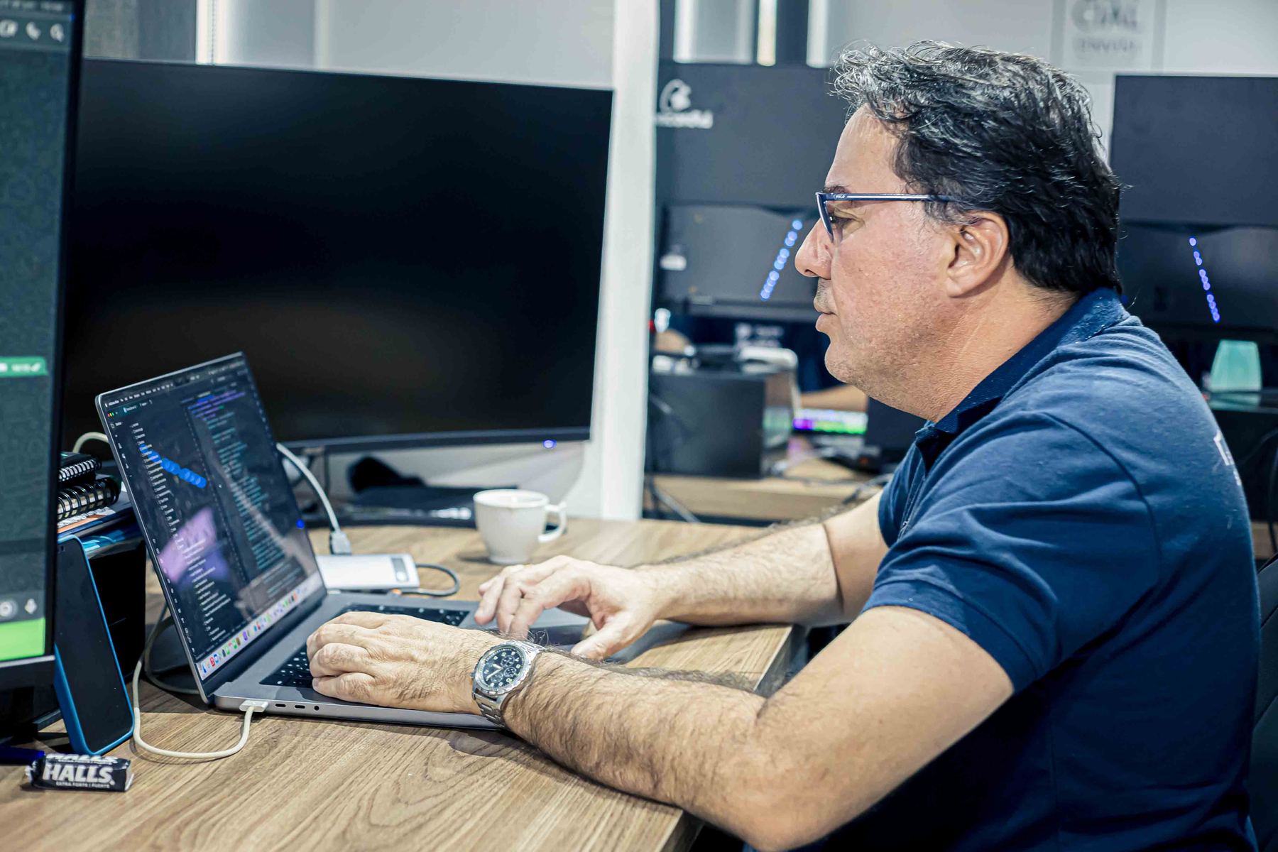 Profissional de óculos programando em um notebook, com monitores ao fundo.