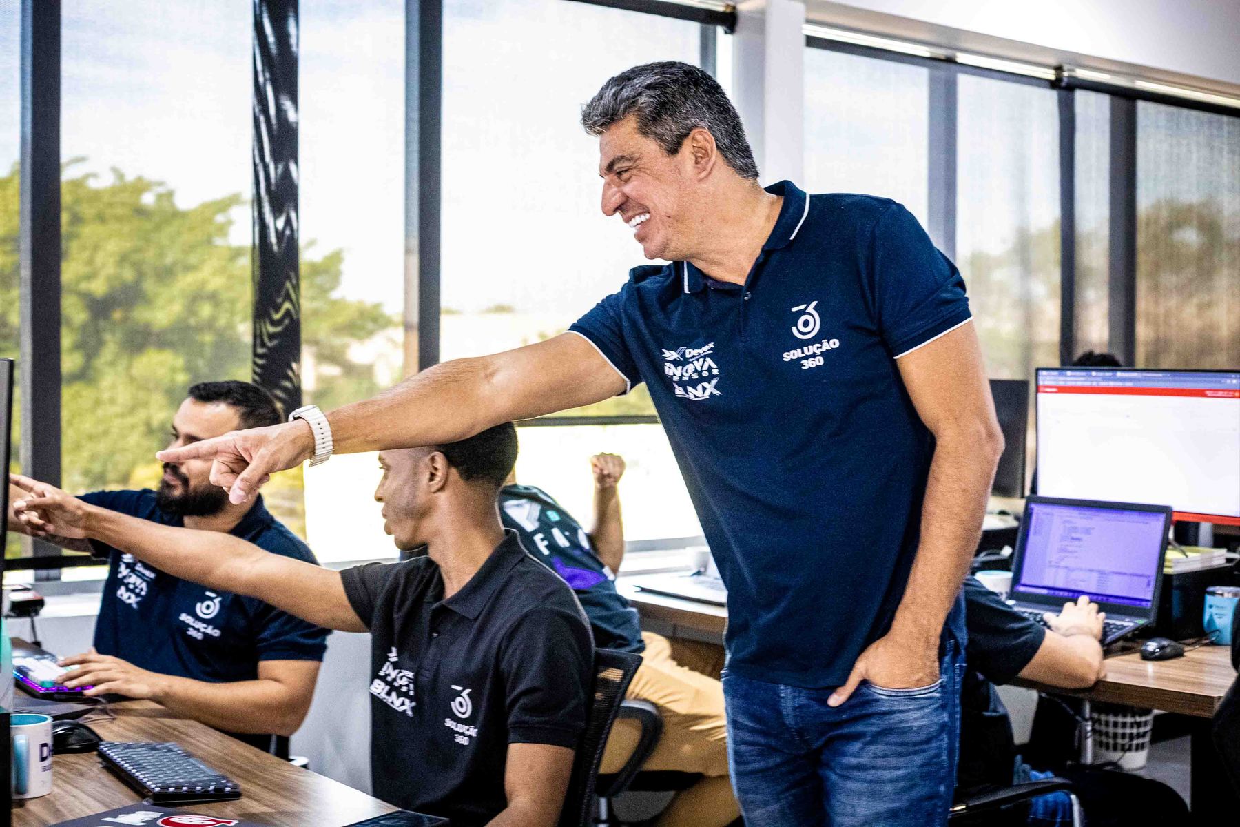 Líder da Devex, de pé, aponta para a tela enquanto orienta colegas sentados à bancada de desenvolvimento