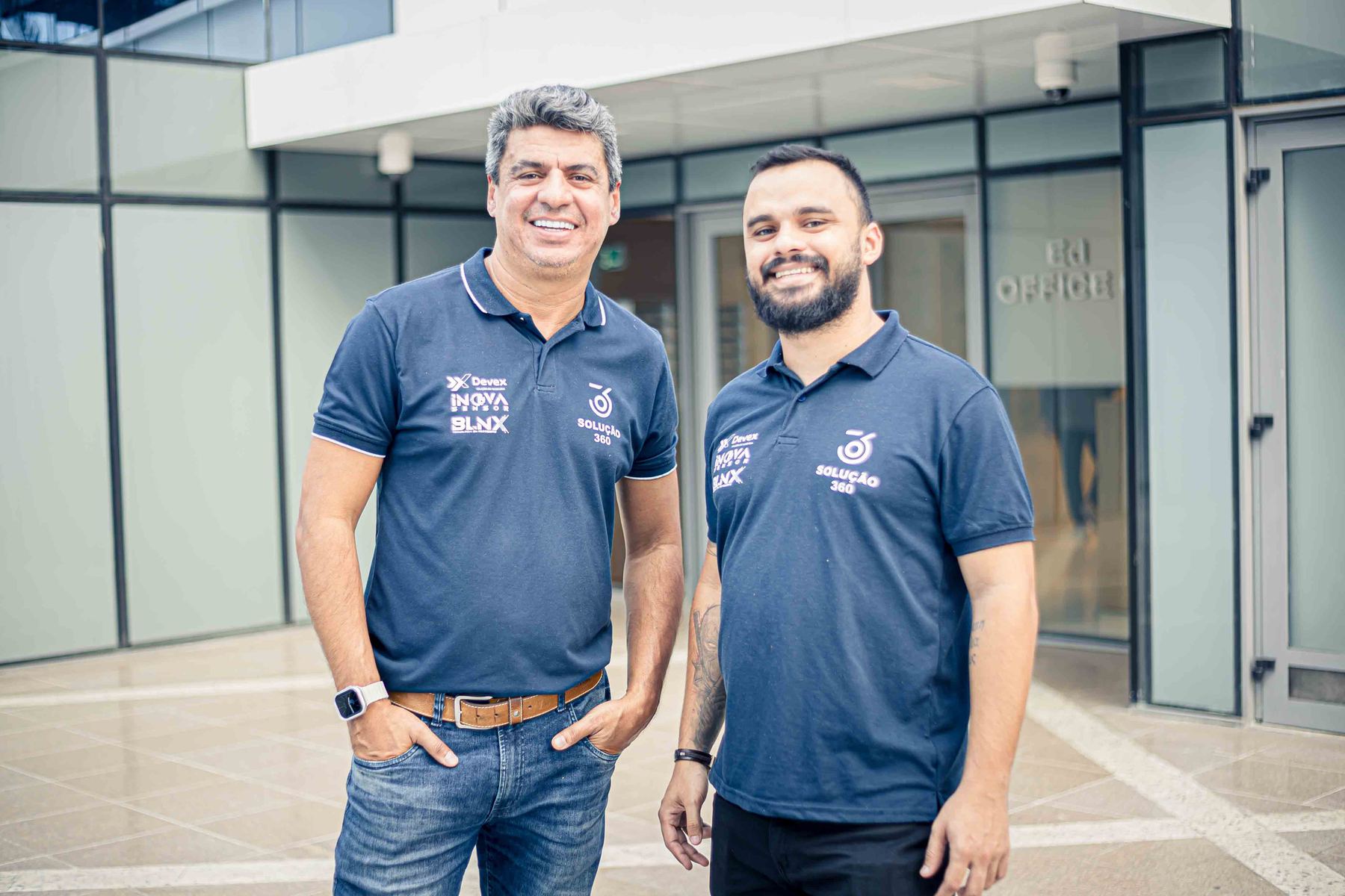 Dois líderes da Devex sorrindo lado a lado na entrada de um prédio comercial.