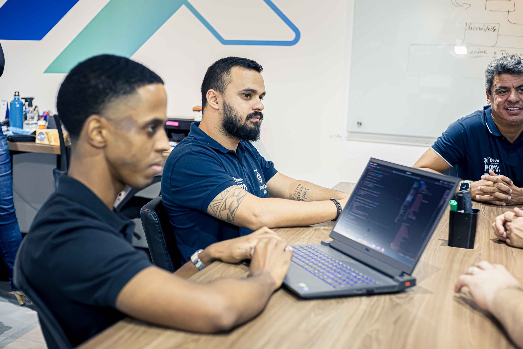 Integrantes do time conversam em volta de uma mesa com notebook aberto, diante de um quadro branco.