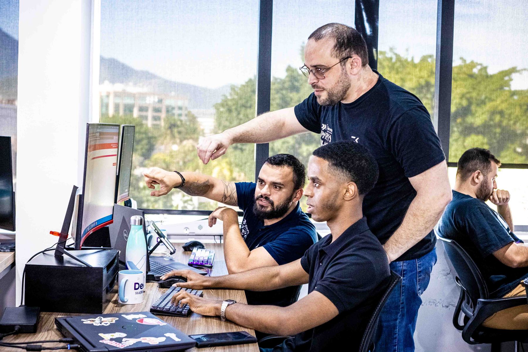 Líder aponta para o monitor orientando dois desenvolvedores, com janela para as montanhas ao fundo.
