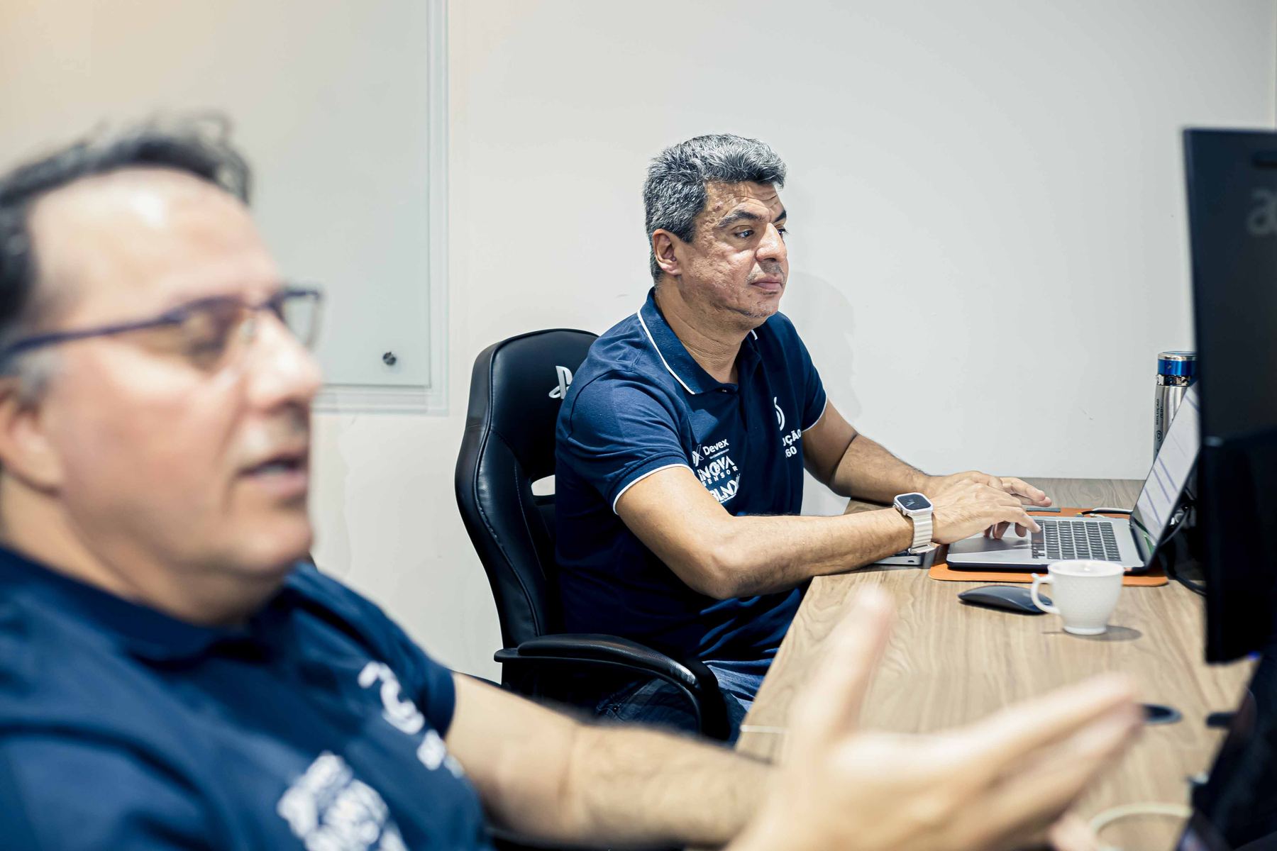 Dois integrantes da Devex conversam à mesa de trabalho, diante de um notebook