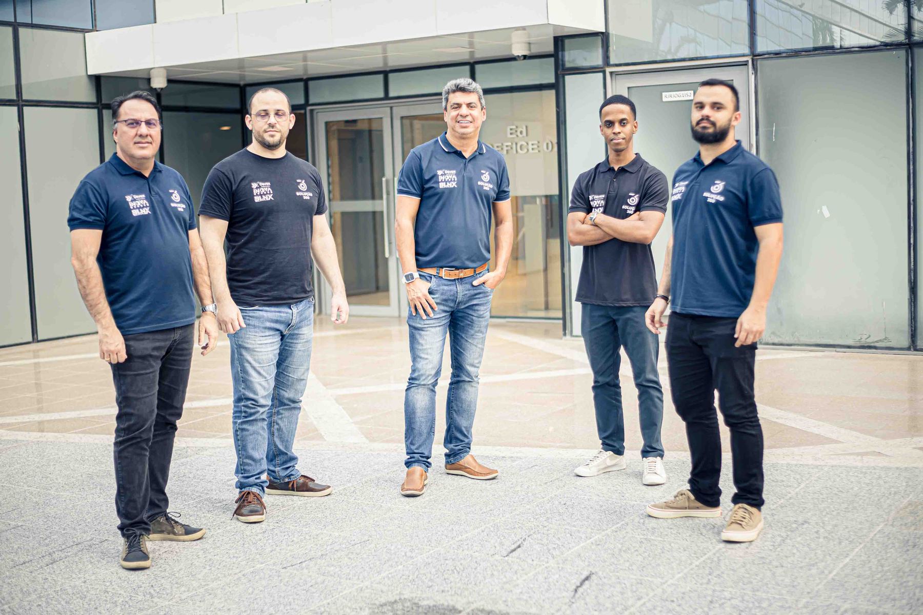 Cinco integrantes do time da Devex, de camisa polo azul-marinho, em frente a um prédio comercial envidraçado.